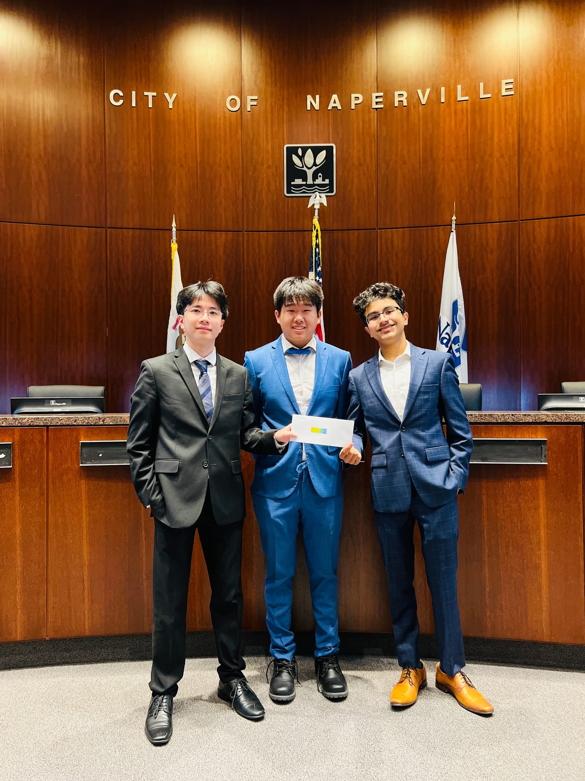 Three BizBuzz team members holding the grant in the Naperville City Council chambers